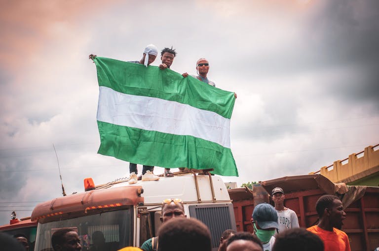 A group of people holding a Nigerian flag, standing on a vehicle in an urban setting, expressing national pride.