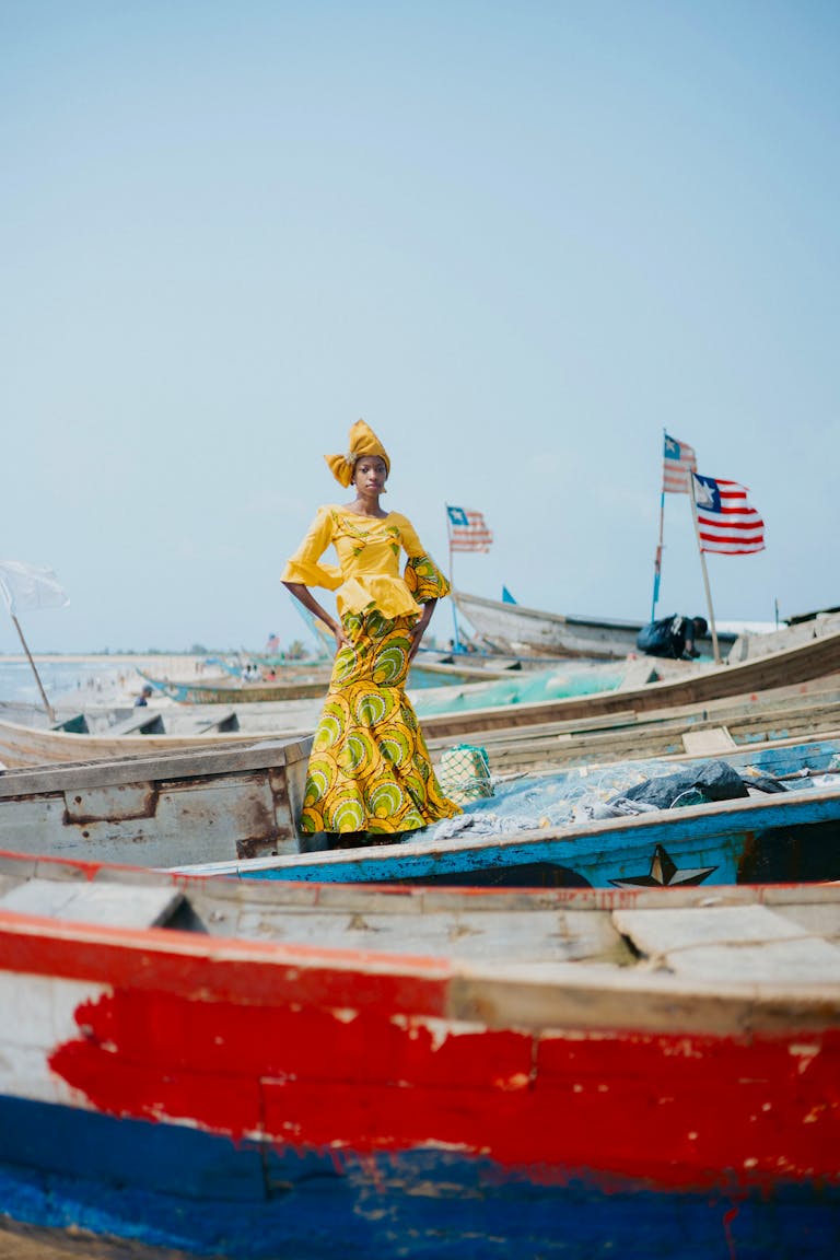 A woman in vibrant traditional attire poses among colorful boats by the sea in Monrovia, Liberia.